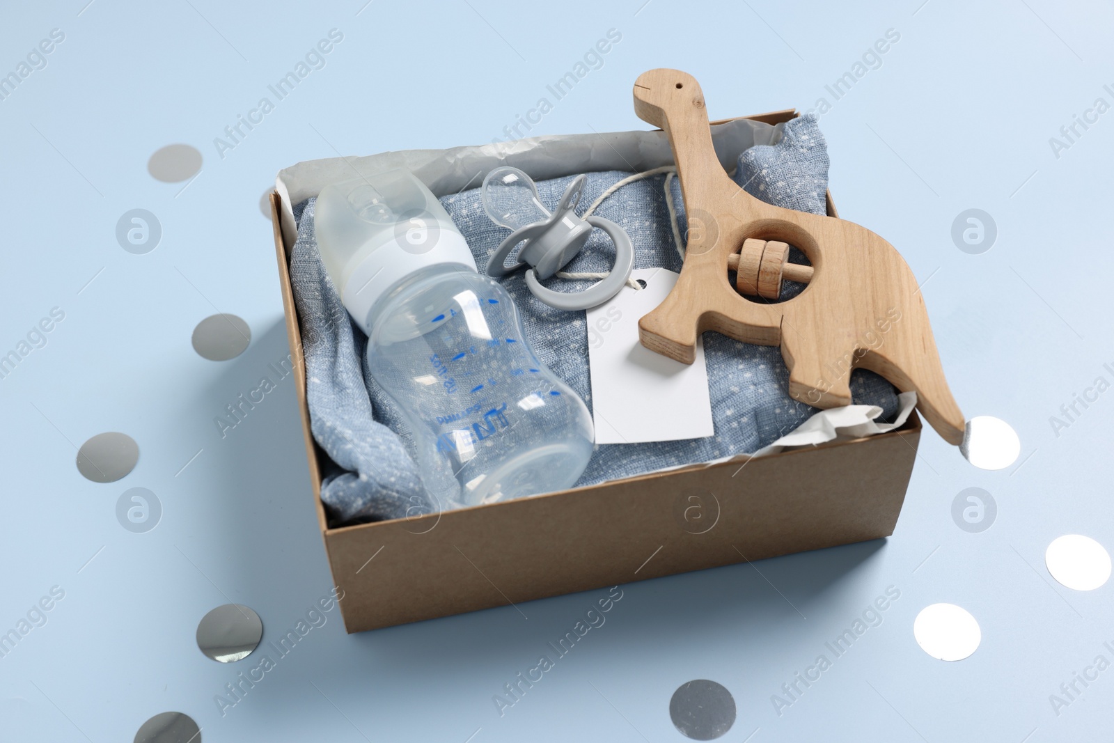 Photo of Different baby accessories with blank card in box and confetti on light blue background