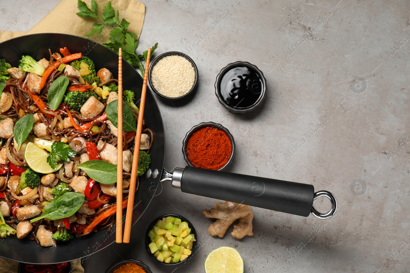 Photo of Stir-fry. Tasty noodles with meat in wok, chopsticks and ingredients on grey textured table, flat lay