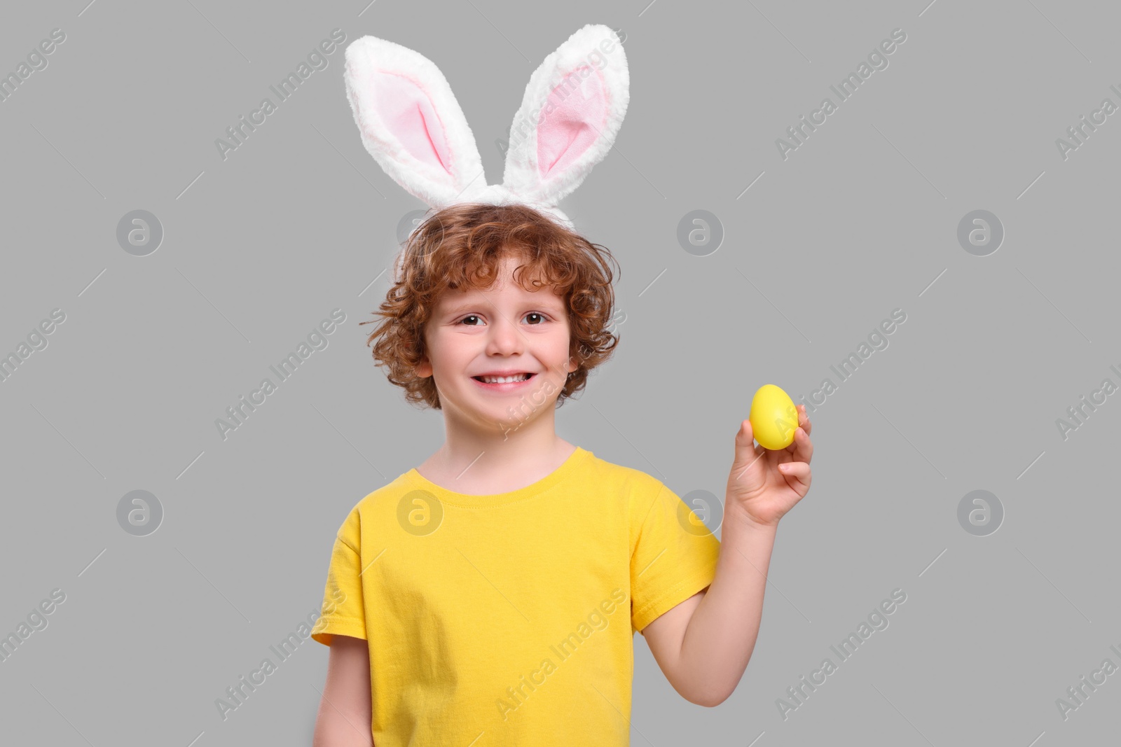 Photo of Portrait of happy boy in cute bunny ears headband holding Easter egg on light grey background