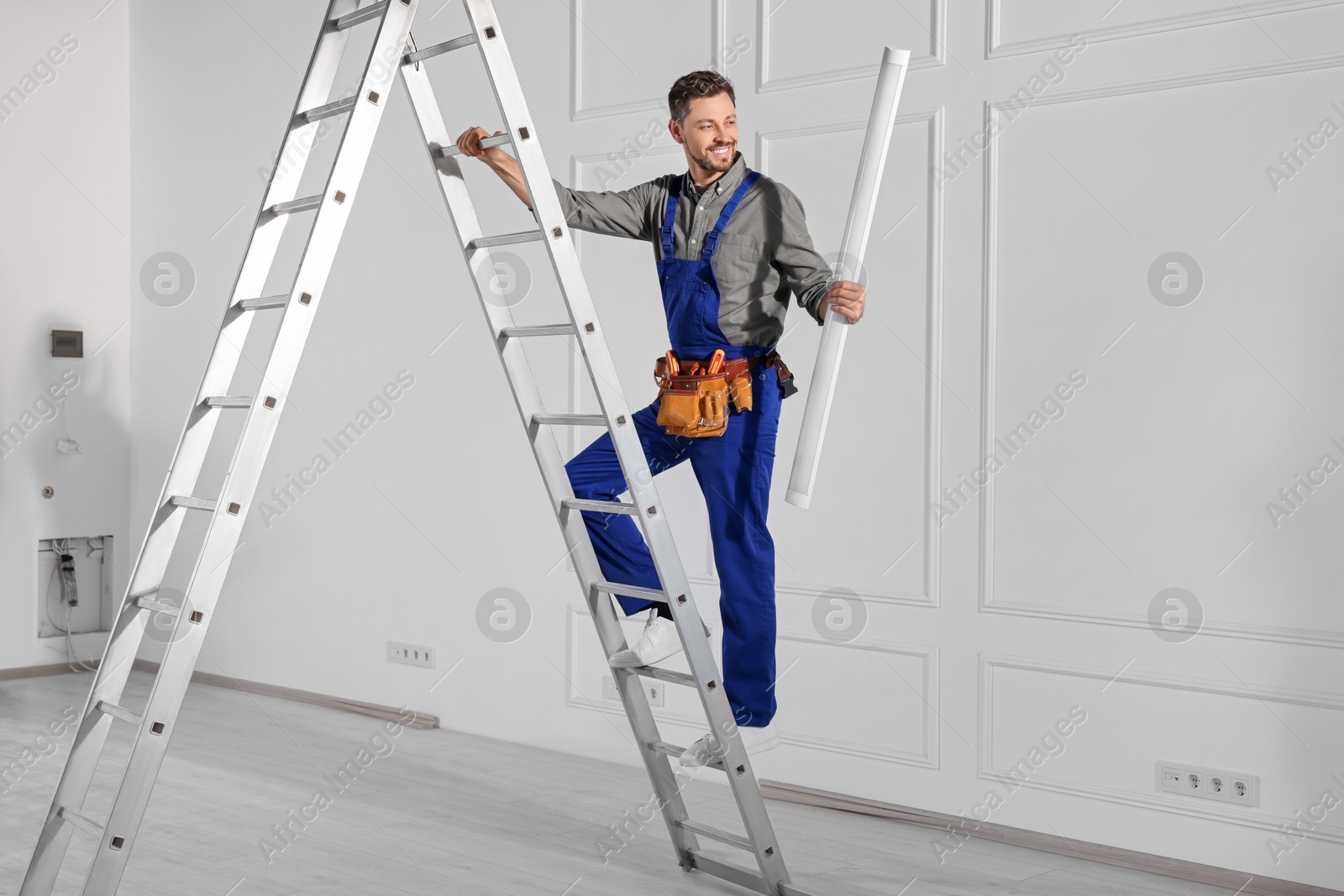 Photo of Electrician in uniform with ceiling lamp on metal ladder indoors