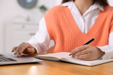 Photo of Young woman writing in notebook while working on laptop at wooden table, closeup