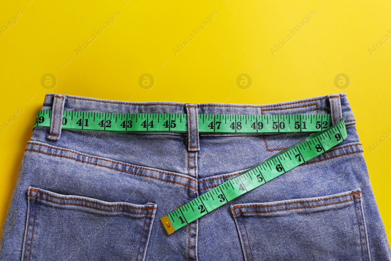 Photo of Jeans with measuring tape on yellow background, top view. Weight loss concept