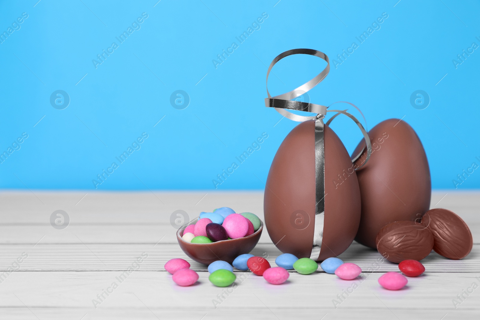 Photo of Delicious chocolate eggs and colorful candies on white wooden table against light blue background. Space for text