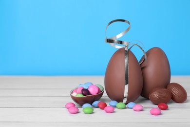 Delicious chocolate eggs and colorful candies on white wooden table against light blue background. Space for text