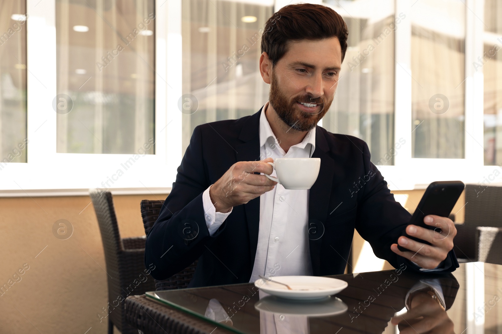 Photo of Handsome bearded man with cup of drink and smartphone at table in cafe