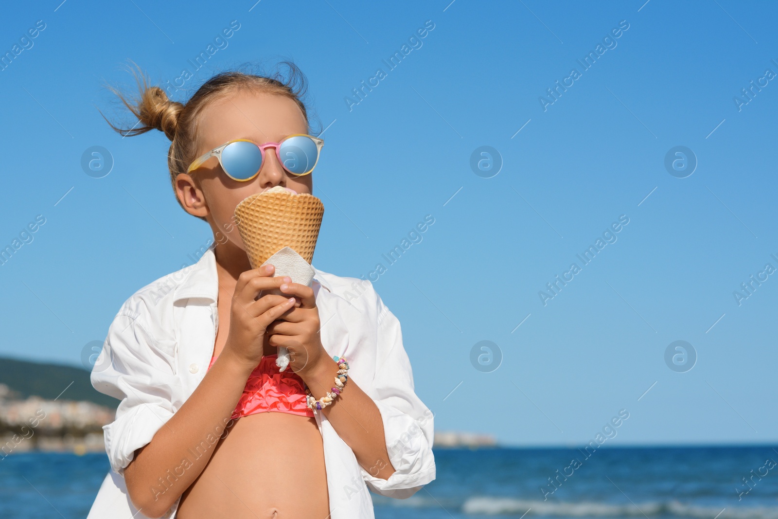 Photo of Adorable little girl eating delicious ice cream near sea on sunny summer day, space for text