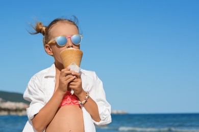 Photo of Adorable little girl eating delicious ice cream near sea on sunny summer day, space for text