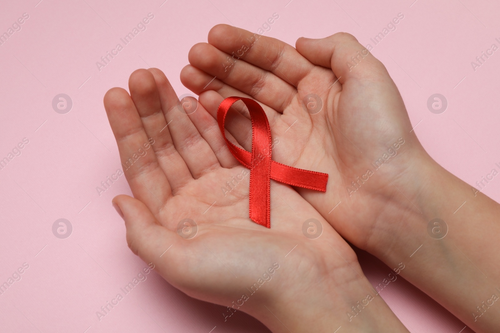 Photo of Little girl holding red ribbon on pink background, closeup. AIDS disease awareness