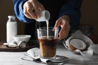 Woman pouring coconut milk into glass of coffee at white wooden table, closeup