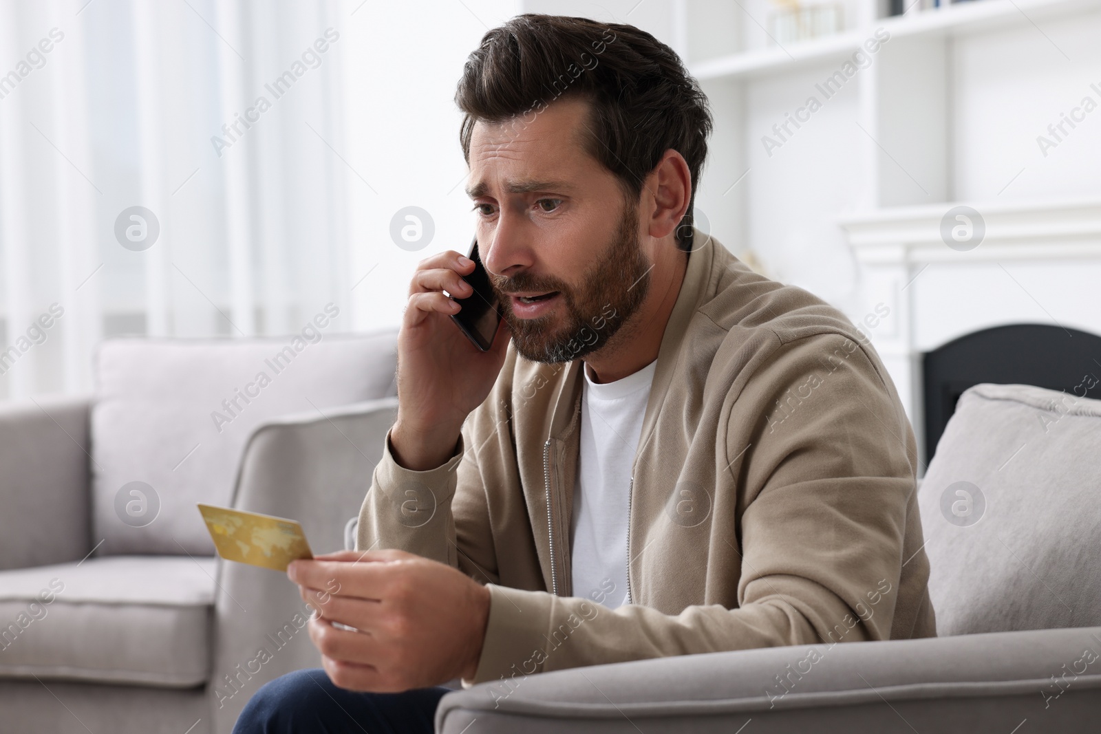 Photo of Upset man with credit card talking on phone in armchair at home. Be careful - fraud