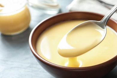 Photo of Spoon of pouring condensed milk over bowl on table, closeup with space for text. Dairy products