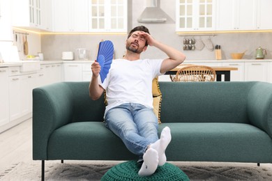 Photo of Bearded man waving blue hand fan to cool himself on sofa at home