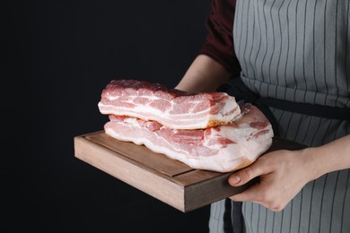 Woman holding wooden board with pieces of raw pork belly on black background, closeup