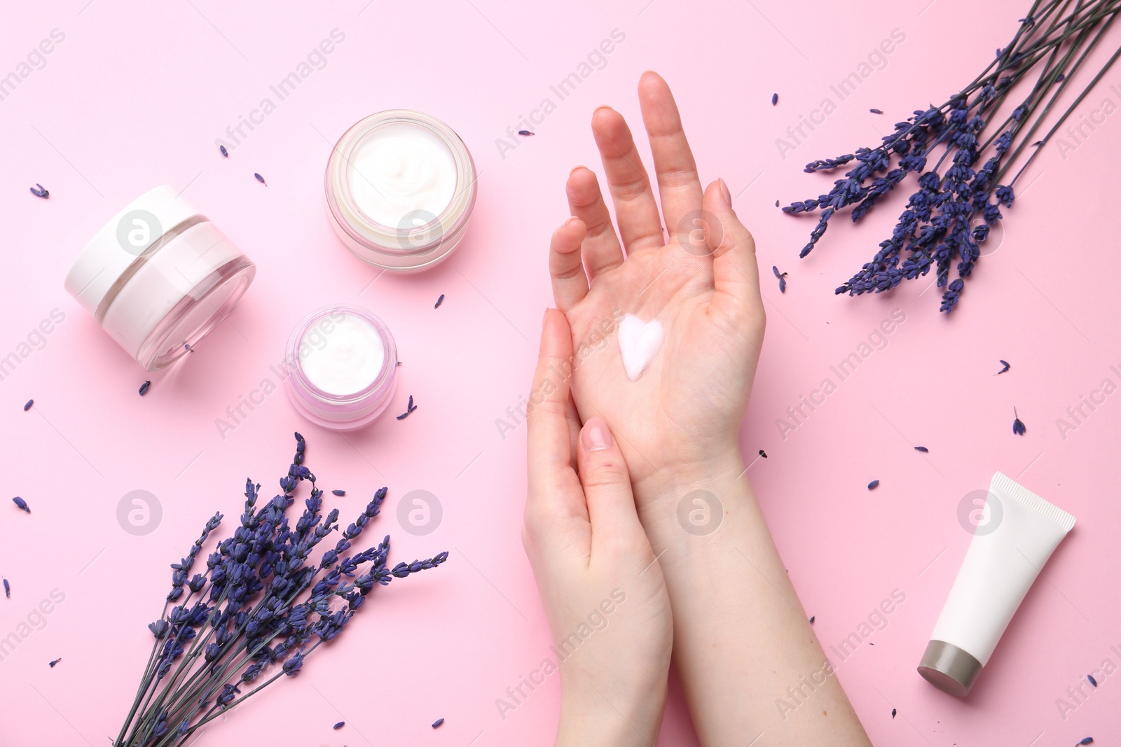 Photo of Woman applying hand cream and lavender flowers on pink background, top view