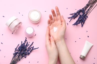 Photo of Woman applying hand cream and lavender flowers on pink background, top view