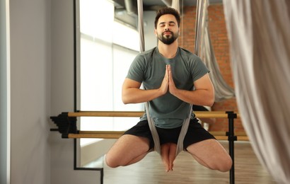 Young man meditating in fly yoga hammock indoors