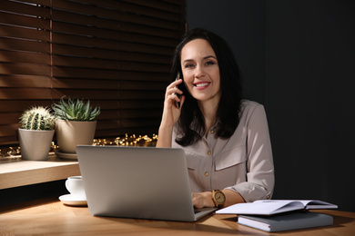 Beautiful mature woman with laptop talking on smartphone at home