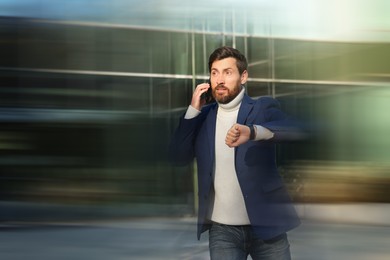 Image of Being late. Worried man talking on phone while checking time and running outdoors. Motion blur effect