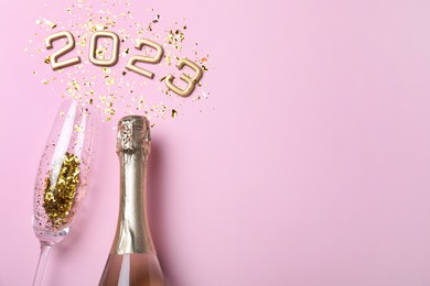 Bottle of sparkling wine, glass, golden confetti and number 2023 on pink background, flat lay with space for text. Happy New Year