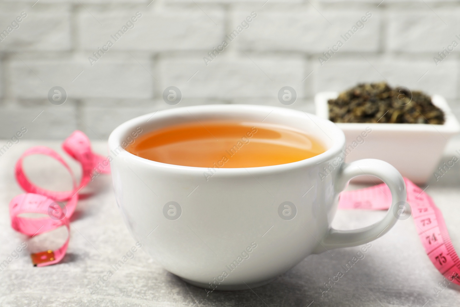 Photo of Cup of herbal diet tea and measuring tape on light grey table against brick wall, closeup. Weight loss concept