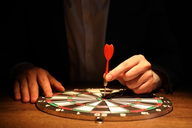 Business targeting concept. Man with dart aiming at dartboard at table in darkness, closeup