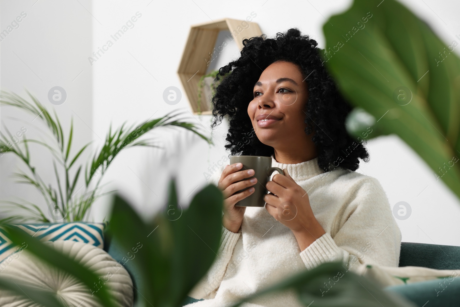 Photo of Relaxing atmosphere. Happy woman with cup of hot drink near beautiful houseplants indoors