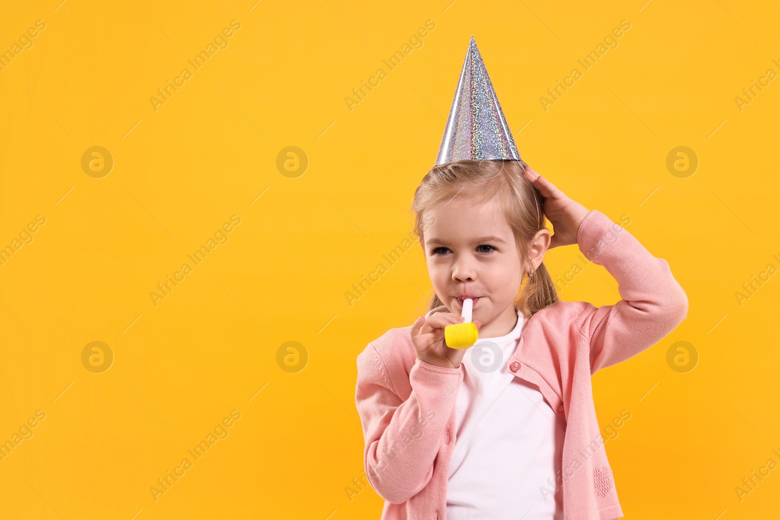 Photo of Birthday celebration. Cute little girl in party hat with blower on orange background, space for text