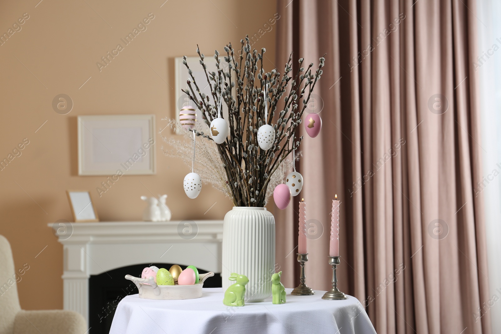 Photo of Pussy willow branches with festively decorated eggs, Easter bunnies and candles on table indoors