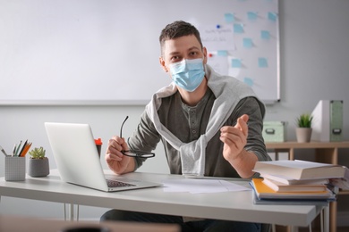 Photo of Teacher with protective mask and laptop sitting at desk in classroom. Reopening after Covid-19 quarantine