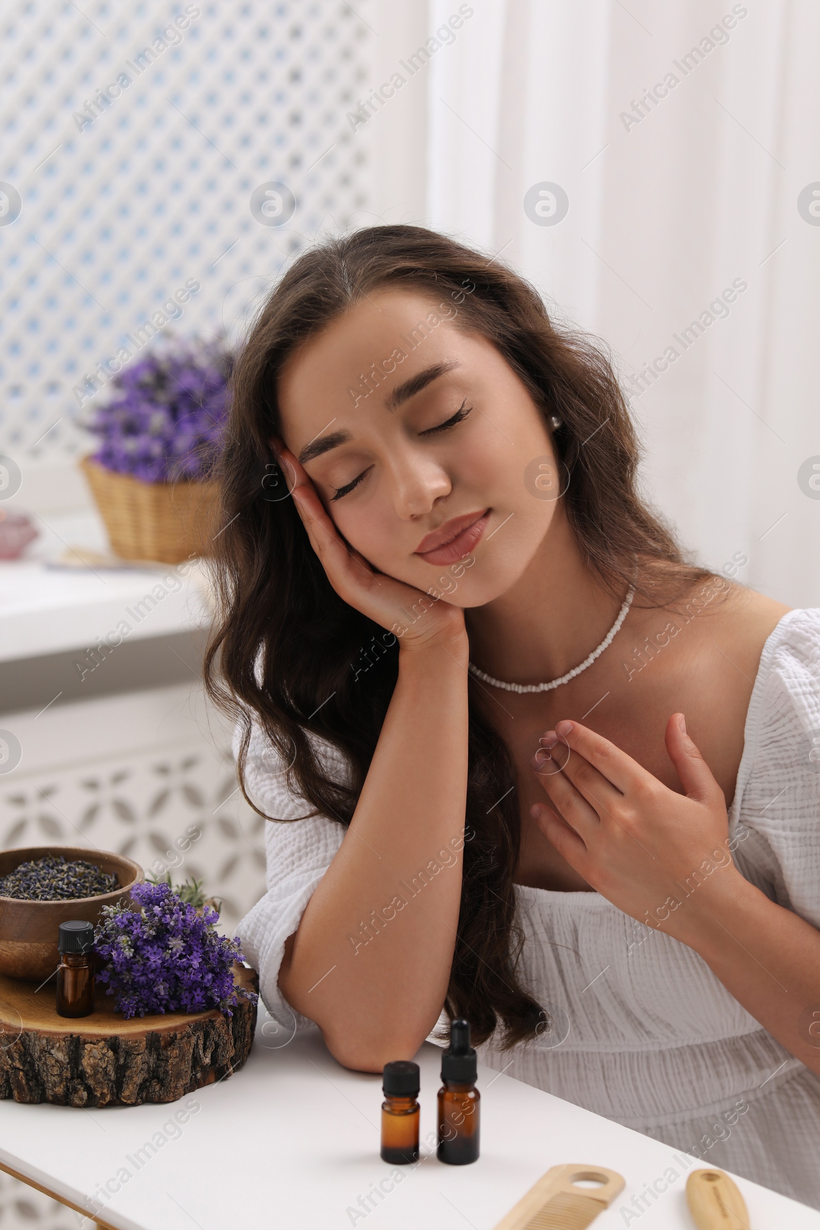 Photo of Beautiful young woman at table with bottles of essential oil and flowers indoors