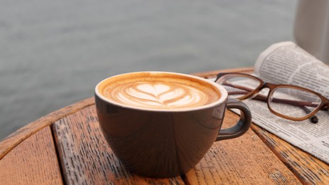 Cup of delicious coffee, eyeglasses and newspaper on wooden table, closeup