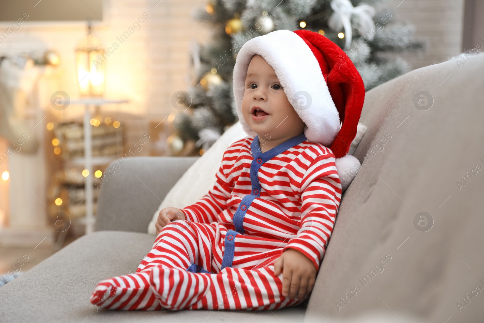 Image of Cute baby in Santa hat sitting on sofa at home. Christmas celebration