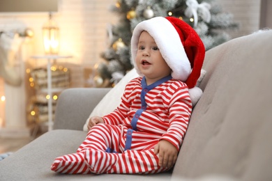 Cute baby in Santa hat sitting on sofa at home. Christmas celebration