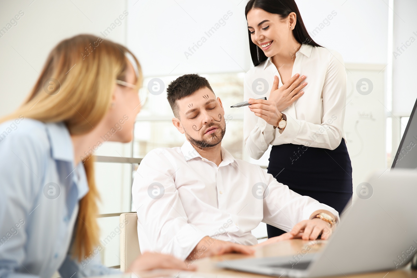 Photo of Young woman drawing on colleague's face while he sleeping in office. Funny joke