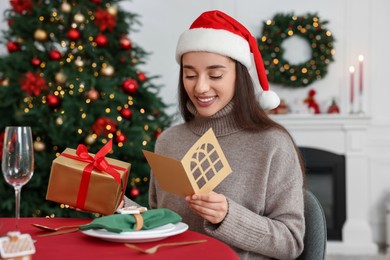 Happy young woman in Santa hat with Christmas gift reading greeting card at served table in decorated room