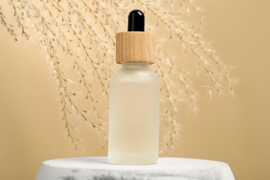 Photo of Bottle of face serum and dried flowers on marble stand against beige background, closeup
