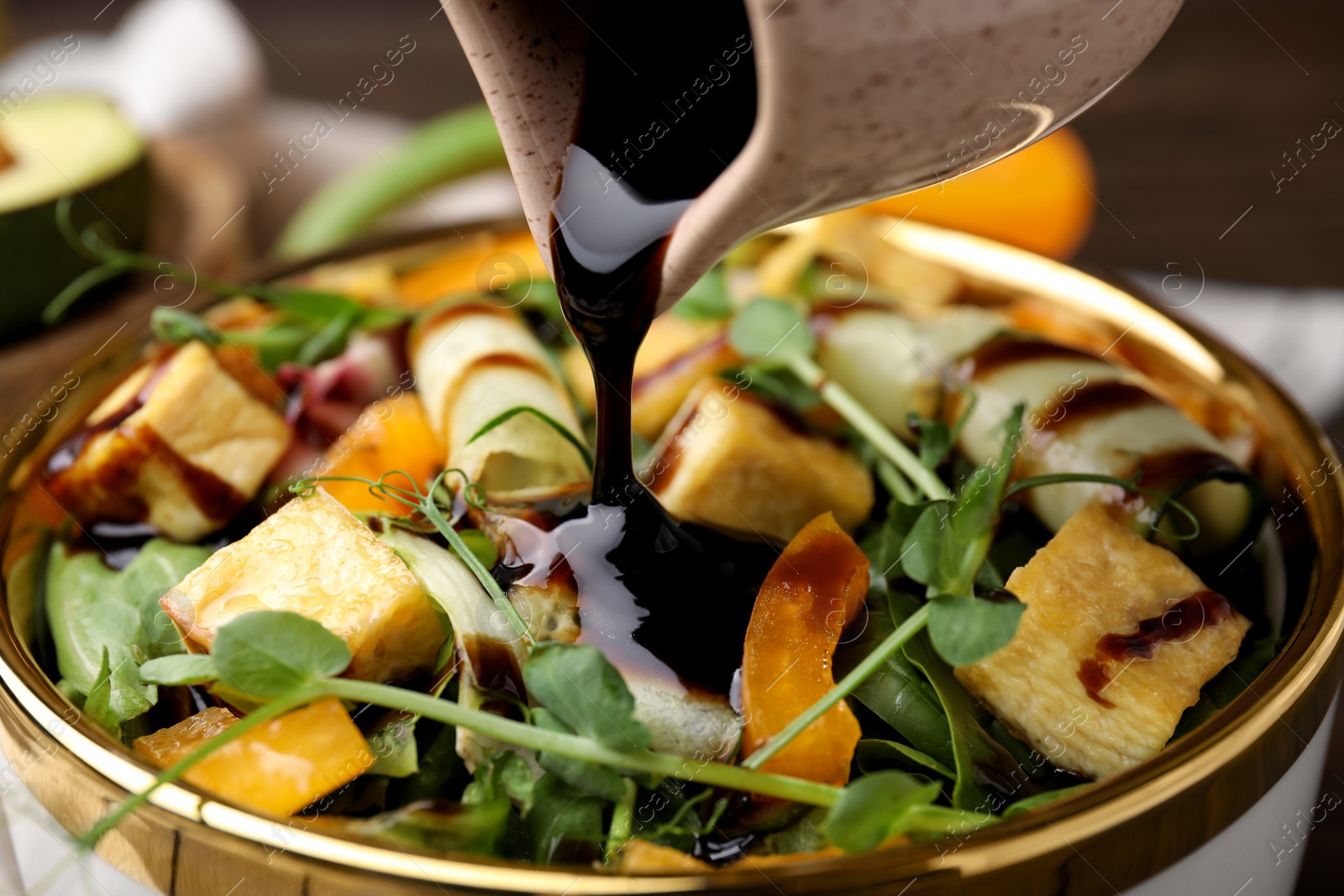 Photo of Pouring balsamic vinegar onto delicious salad with tofu and vegetables in bowl, closeup