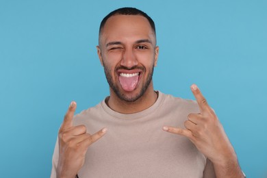 Photo of Happy young man showing his tongue and rock gesture on light blue background