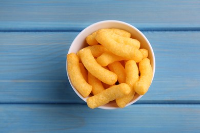 Photo of Bowl of tasty cheesy corn puffs on blue wooden table, top view