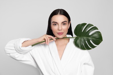 Photo of Woman in bathrobe holding leaf of monstera on light grey background. Spa treatment