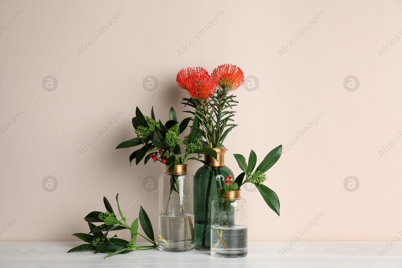 Photo of Vases with beautiful flowers and branches on white wooden table near beige wall