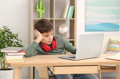 Photo of Tired boy with red headphones using laptop at desk in room. Home workplace