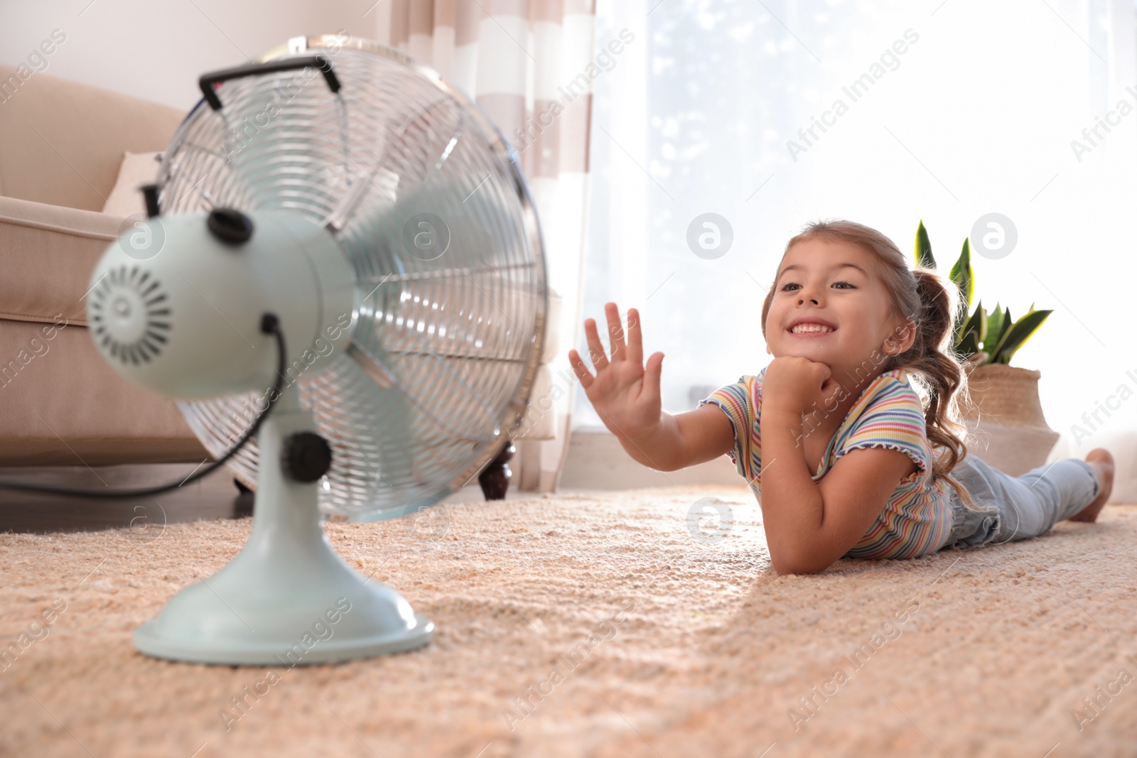 Photo of Little girl enjoying air flow from fan on floor in living room. Summer heat