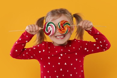 Happy girl covering eyes with lollipops on orange background