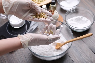 Photo of Woman in gloves filling bath bomb mold with dried flower buds at wooden table, closeup