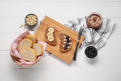 Photo of Toasts with tasty nut butter, banana slices, blueberries and peanuts on white wooden table, flat lay