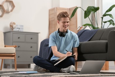 Photo of Online learning. Smiling teenage boy writing essay near laptop at home