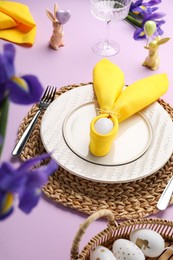 Photo of Festive table setting with painted eggs, plates and iris flowers on lilac background. Easter celebration