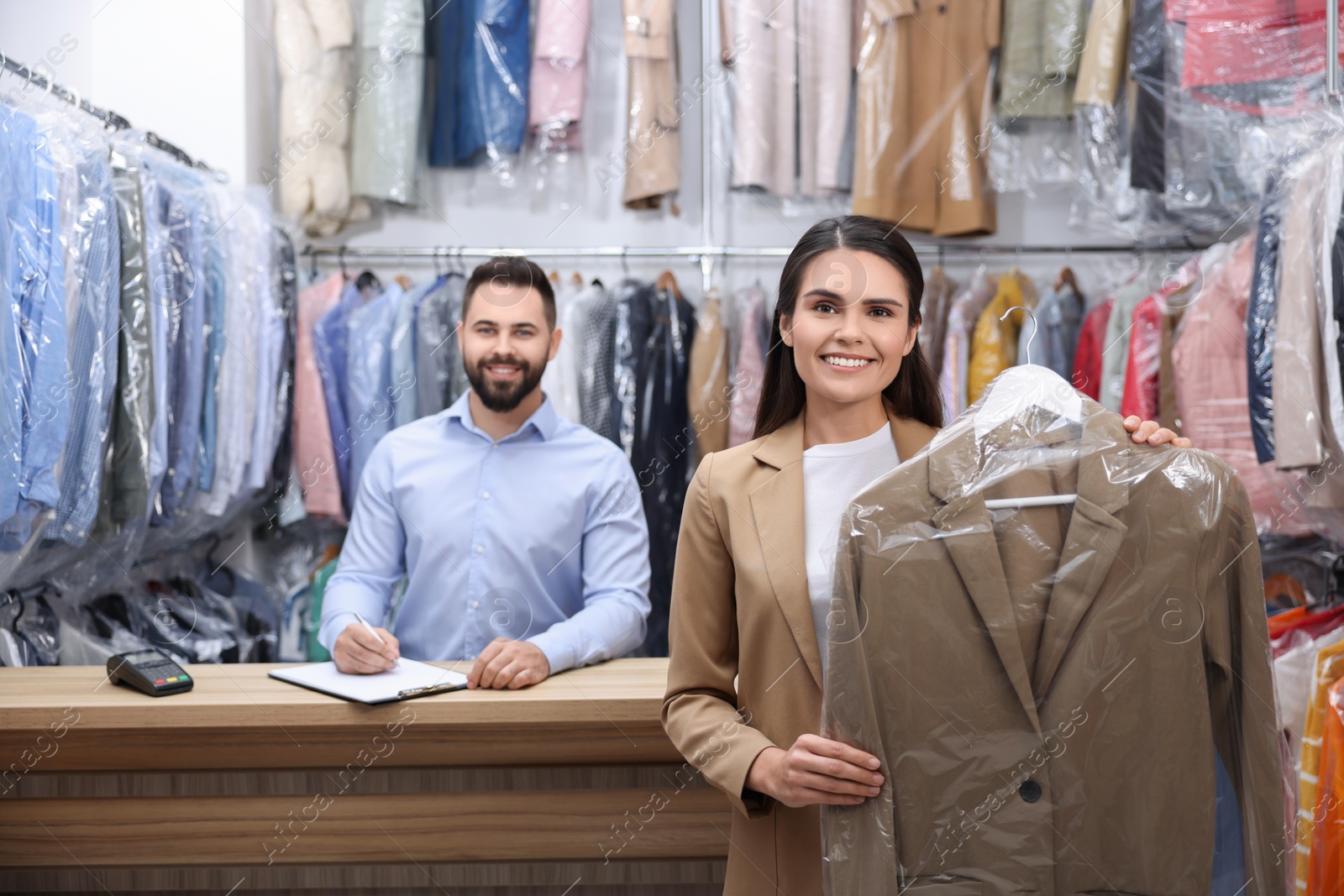 Photo of Dry-cleaning service. Happy woman holding hanger with coat indoors. Worker taking notes at workplace