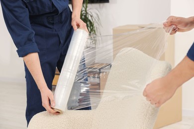 Photo of Male movers with stretch film wrapping armchair in house, closeup